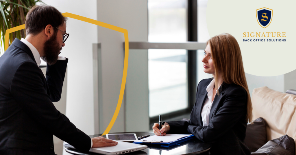 Standing male employee speaking to female employee seated at table with clipboard, laptop, and tablet